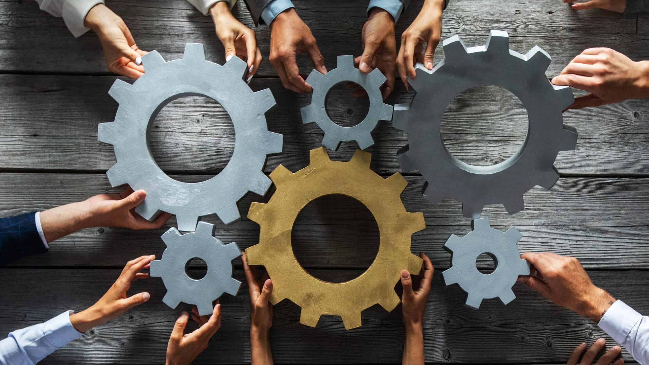 Group of business people joining together silver and golden coloured gears on table at workplace.