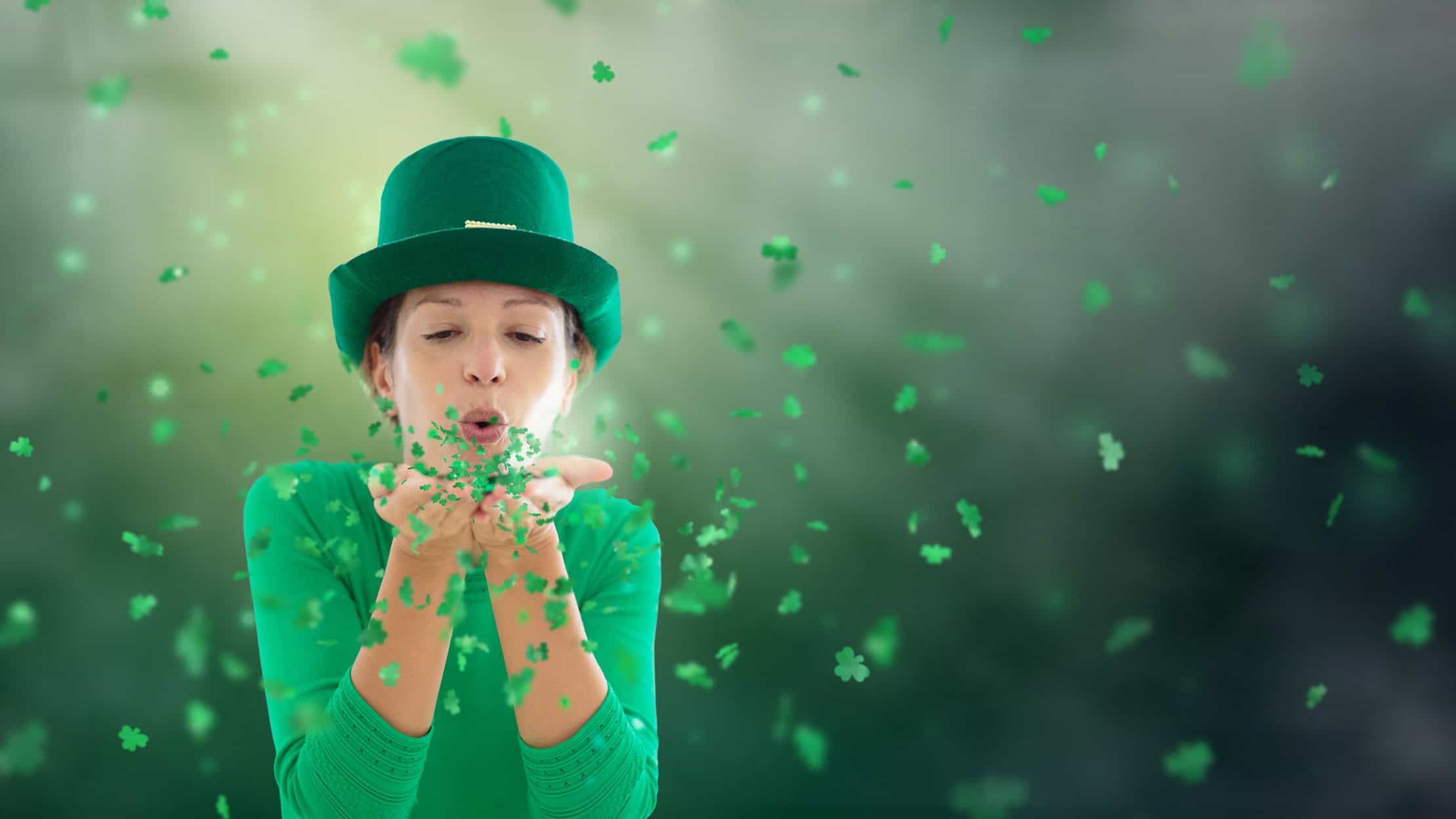 Woman in green leprechaun hat blowing shamrock confetti.