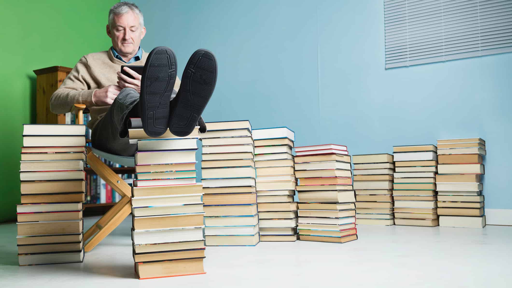 Man reading an e-book with his feet up and piles of books next to him.