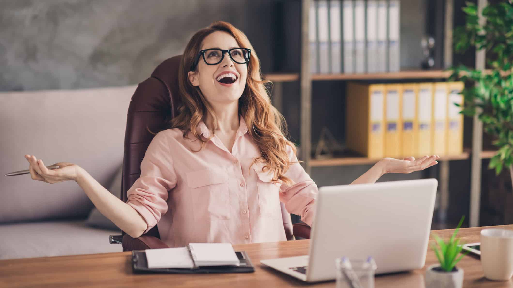 Woman with an amazed expression has her hands and arms out with a laptop in front of her.