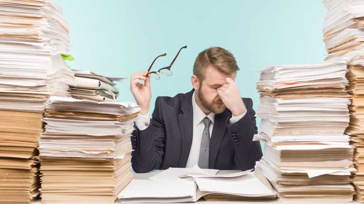 Stacks of files and folders next to businessman who is stressed.