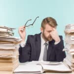 Stacks of files and folders next to businessman who is stressed.