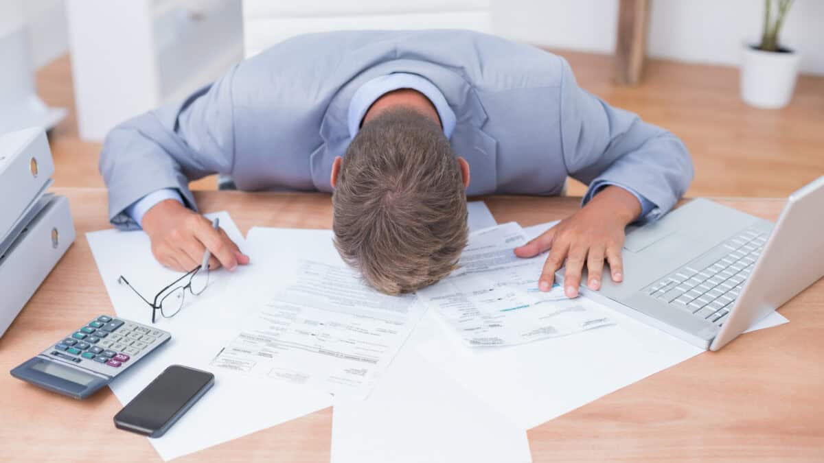 Devastated man with his head on his office desk with paperwork and a laptop.