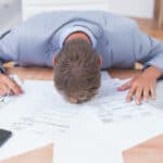 Devastated man with his head on his office desk with paperwork and a laptop.