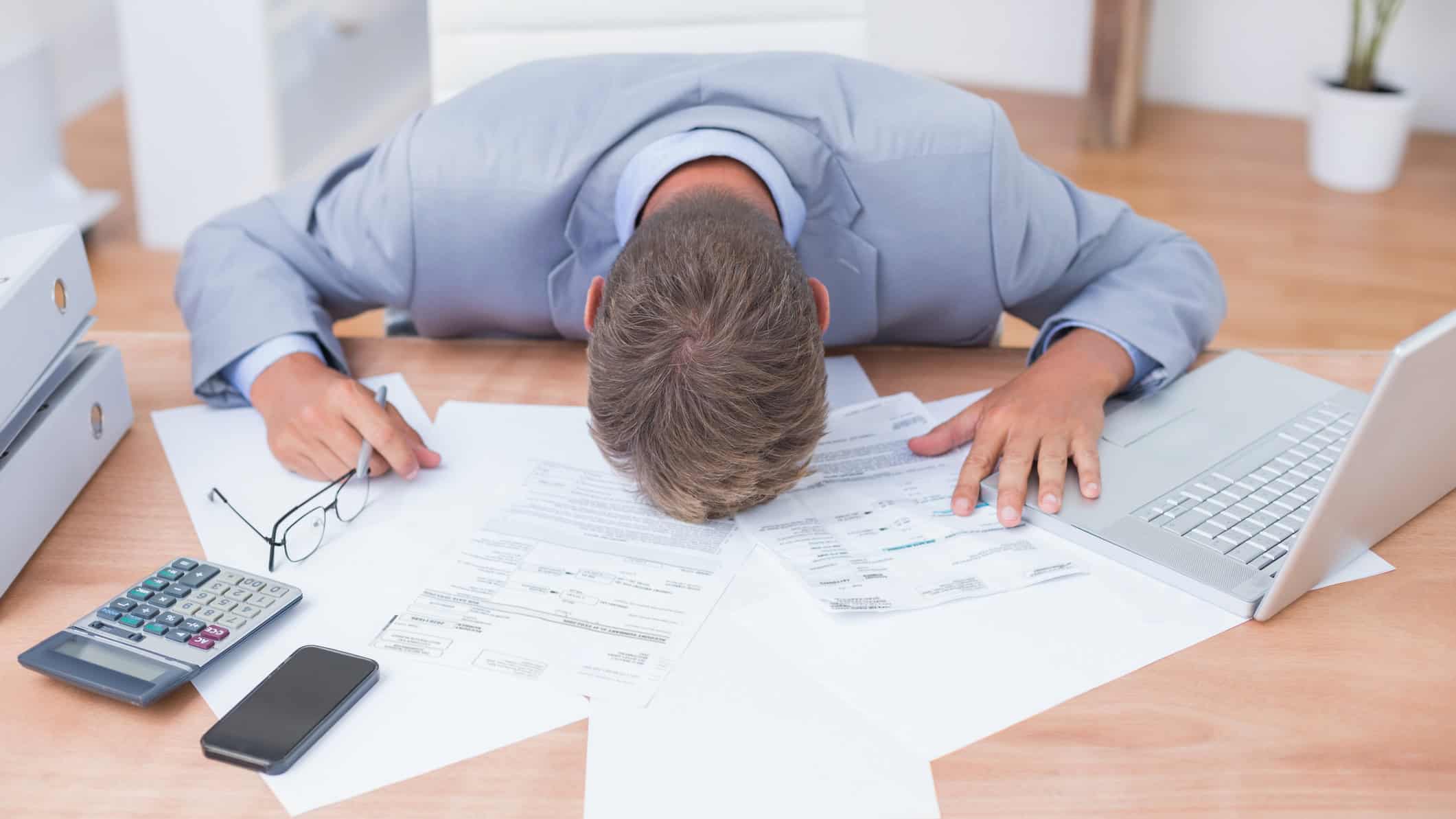 Devastated man with his head on his office desk with paperwork and a laptop.