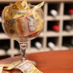 A large clear wine glass on the left of the image filled with fifty dollar notes on a timber table with a wine cellar or cabinet with bottles in the background.