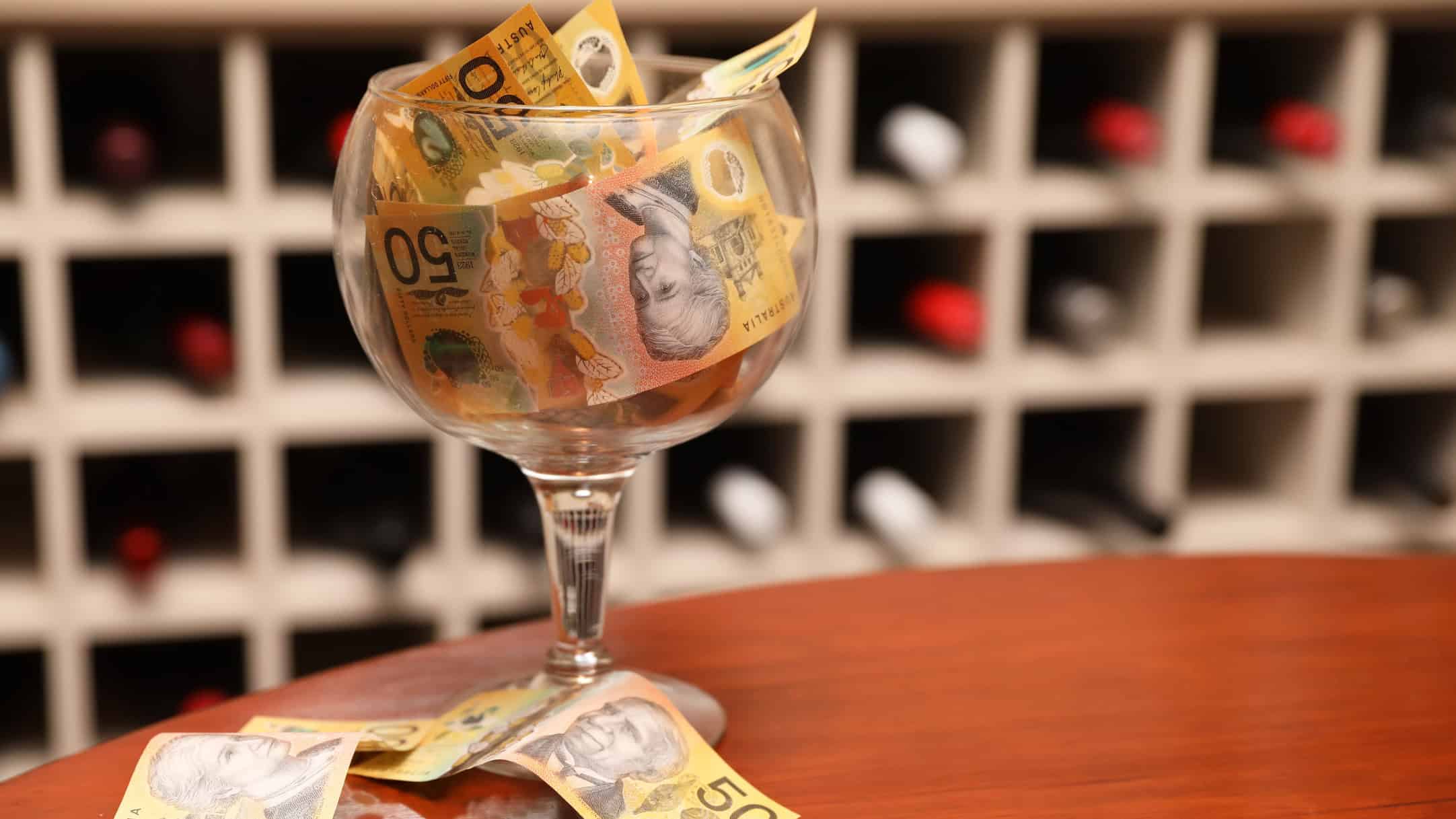 A large clear wine glass on the left of the image filled with fifty dollar notes on a timber table with a wine cellar or cabinet with bottles in the background.