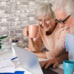 Elderly couple using laptop at home while drinking a cup of coffee.