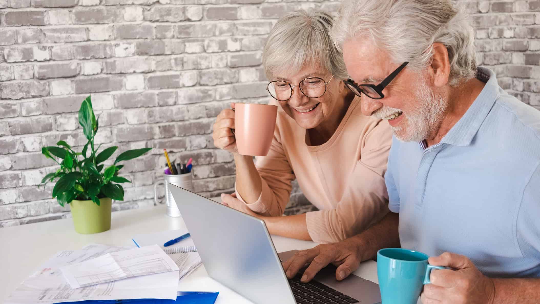 Elderly couple using laptop at home while drinking a cup of coffee.