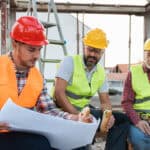 Three people at a building site discussing a plan whilst eating.