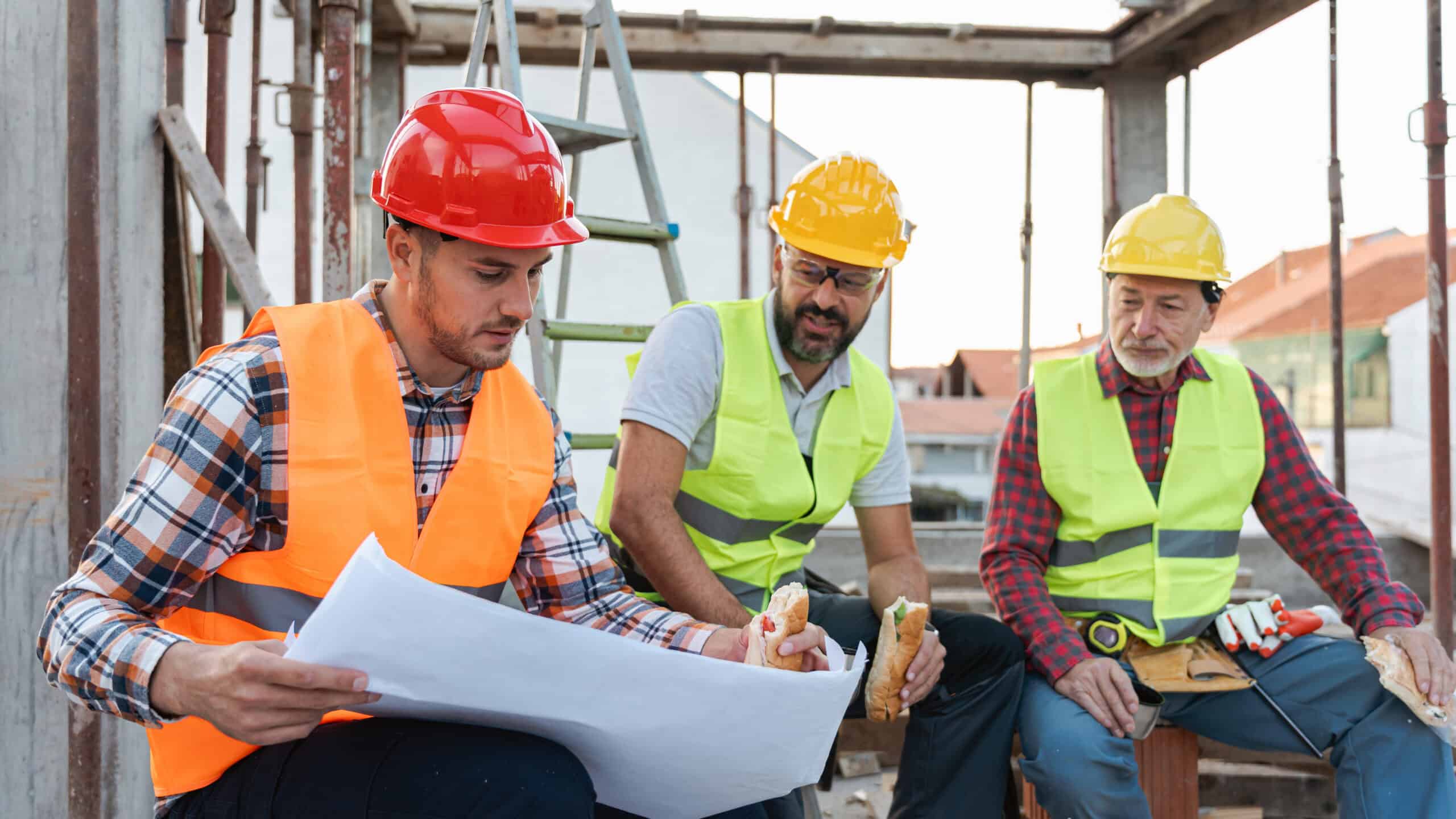 Three people at a building site discussing a plan whilst eating.