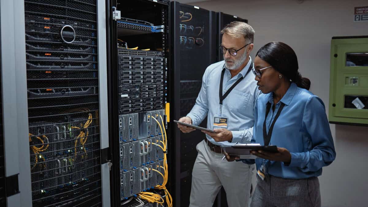 Man and woman holding a tablet and clipboard while looking at the backside of server racks in a data centre.