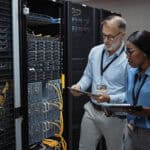 Man and woman holding a tablet and clipboard while looking at the backside of server racks in a data centre.