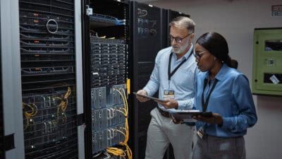 Man and woman holding a tablet and clipboard while looking at the backside of server racks in a data centre.