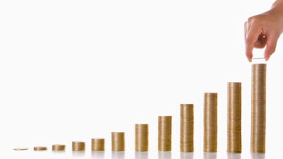 A hand is adding more coins to the highest stack, with twelve stacks of coins showing growth every month for a year.