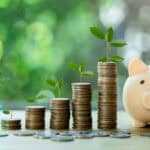 Stacks of coins in ascending order with a plant on top, next to a piggy bank.