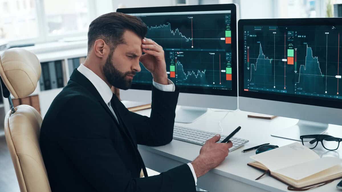 Stressed man in an an office with his eyes closed and phone in his hand, with investing graphs open on two iMacs.