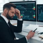 Stressed man in an an office with his eyes closed and phone in his hand, with investing graphs open on two iMacs.
