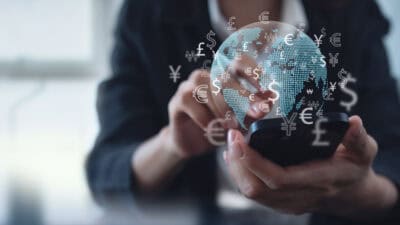 Woman using mobile phone for digital banking, with hologram of globe and different currency symbols.