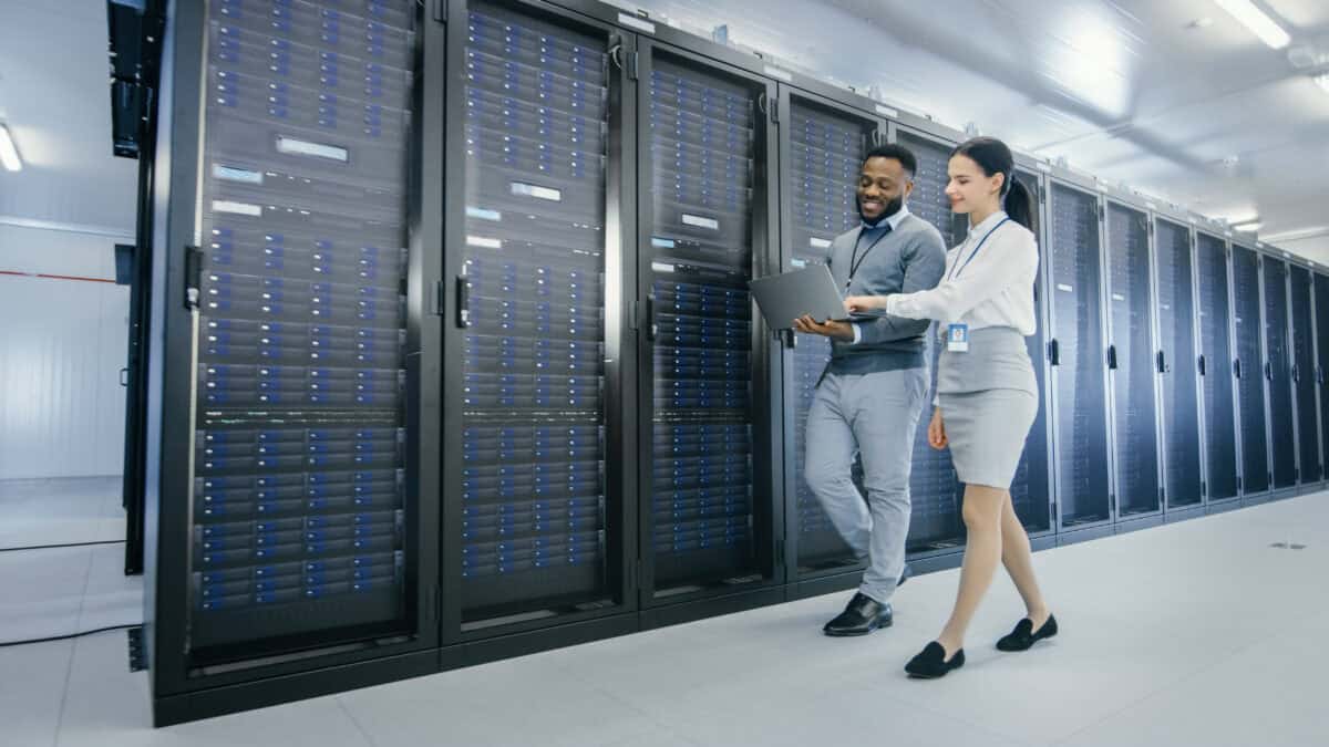 IT technician and young female intern walking next to sever racks in a data centre while using a laptop.