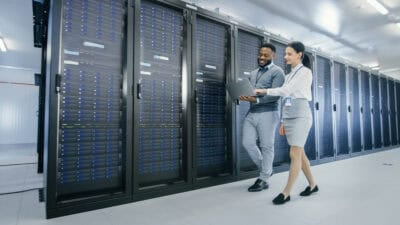 IT technician and young female intern walking next to sever racks in a data centre while using a laptop.