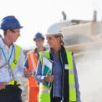 Two people wearing hard hats talking with each other at a mine site, with two workers in the background.