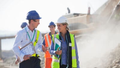 Two people wearing hard hats talking with each other at a mine site, with two workers in the background.