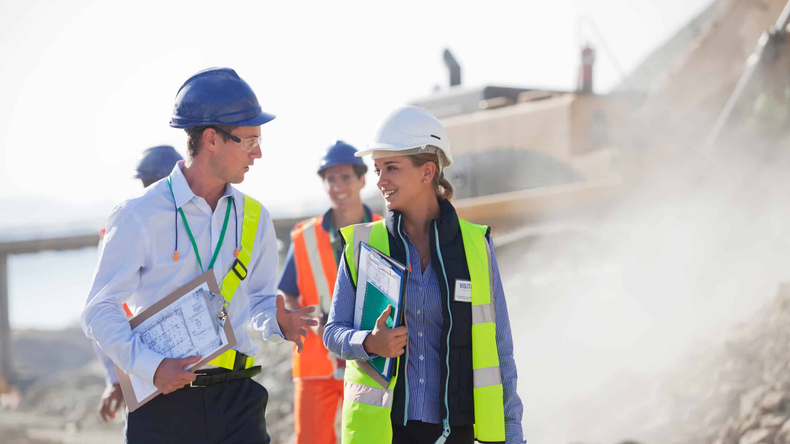Two people wearing hard hats talking with each other at a mine site, with two workers in the background.