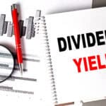 Dividend yield written on a notebook with a chart, pen, and magnifying glass next to it.