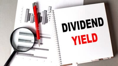 Dividend yield written on a notebook with a chart, pen, and magnifying glass next to it.