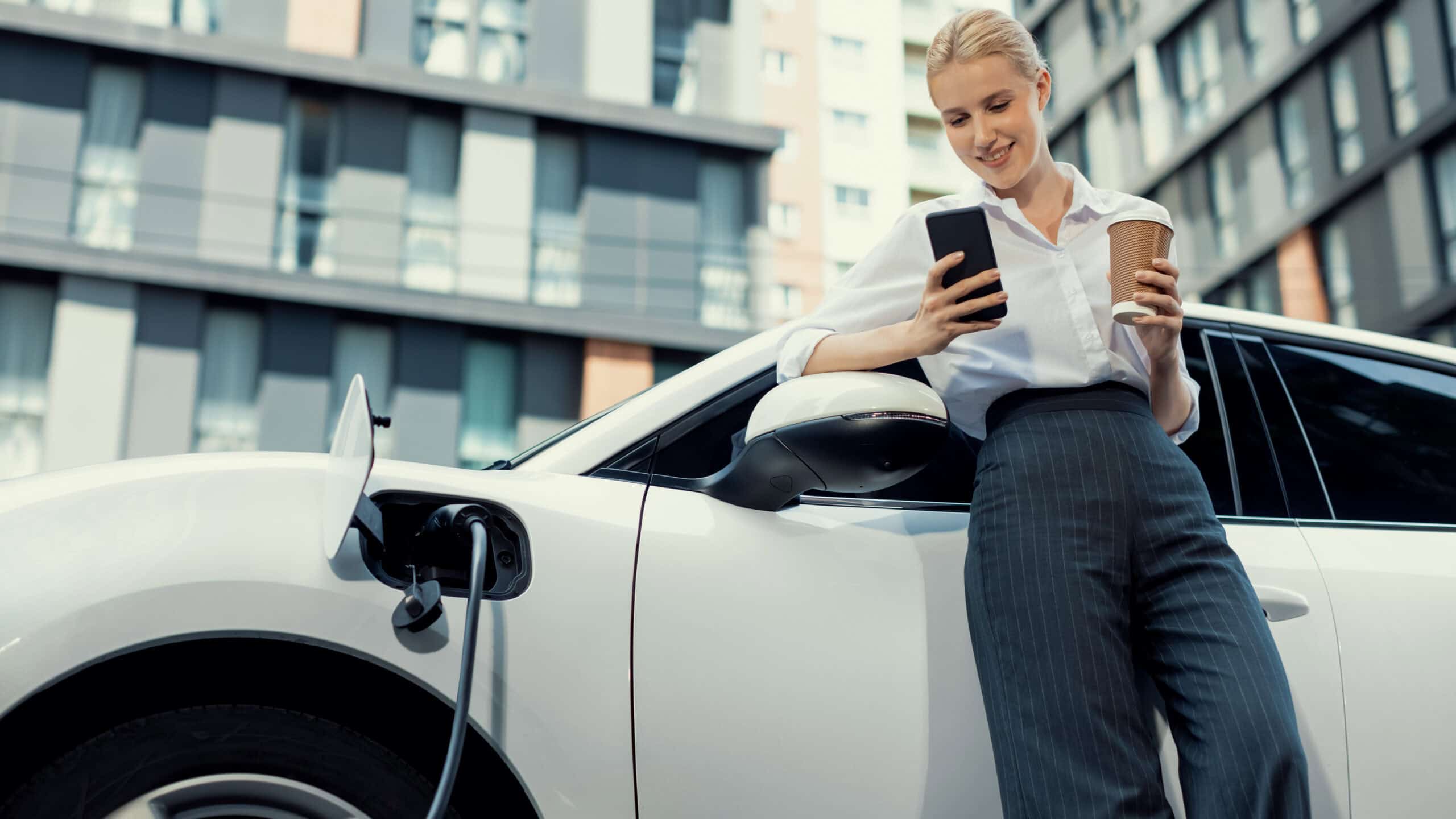 Smiling woman with a coffee in hand using a smartphone while her electric vehicle charges.
