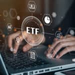 Person working on a computer with a hologram of the word ETF along with finance-related images.