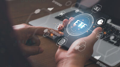 Man holding a smartphone with a hologram of the word ETF along with finance-related images.