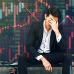 Stressed businessman sits in panic amid digital stock market financial background.