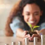 Happy girl holding a plant and soil in front of ascending piles of coins.