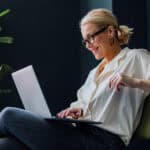 Cheerful smiling businesswoman sitting on a chair and typing business report on a laptop keyboard.