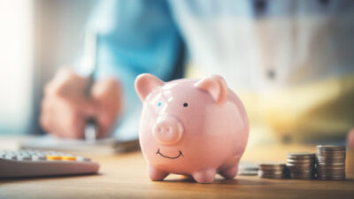 Person writing notes with a piggy bank, calculator, and an ascending pile of coins on the table.