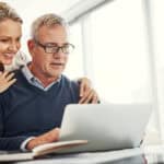 Happy wife holding her hands on her husband's shoulders while both look at a laptop.