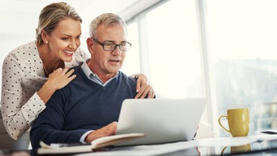 Happy wife holding her hands on her husband's shoulders while both look at a laptop.