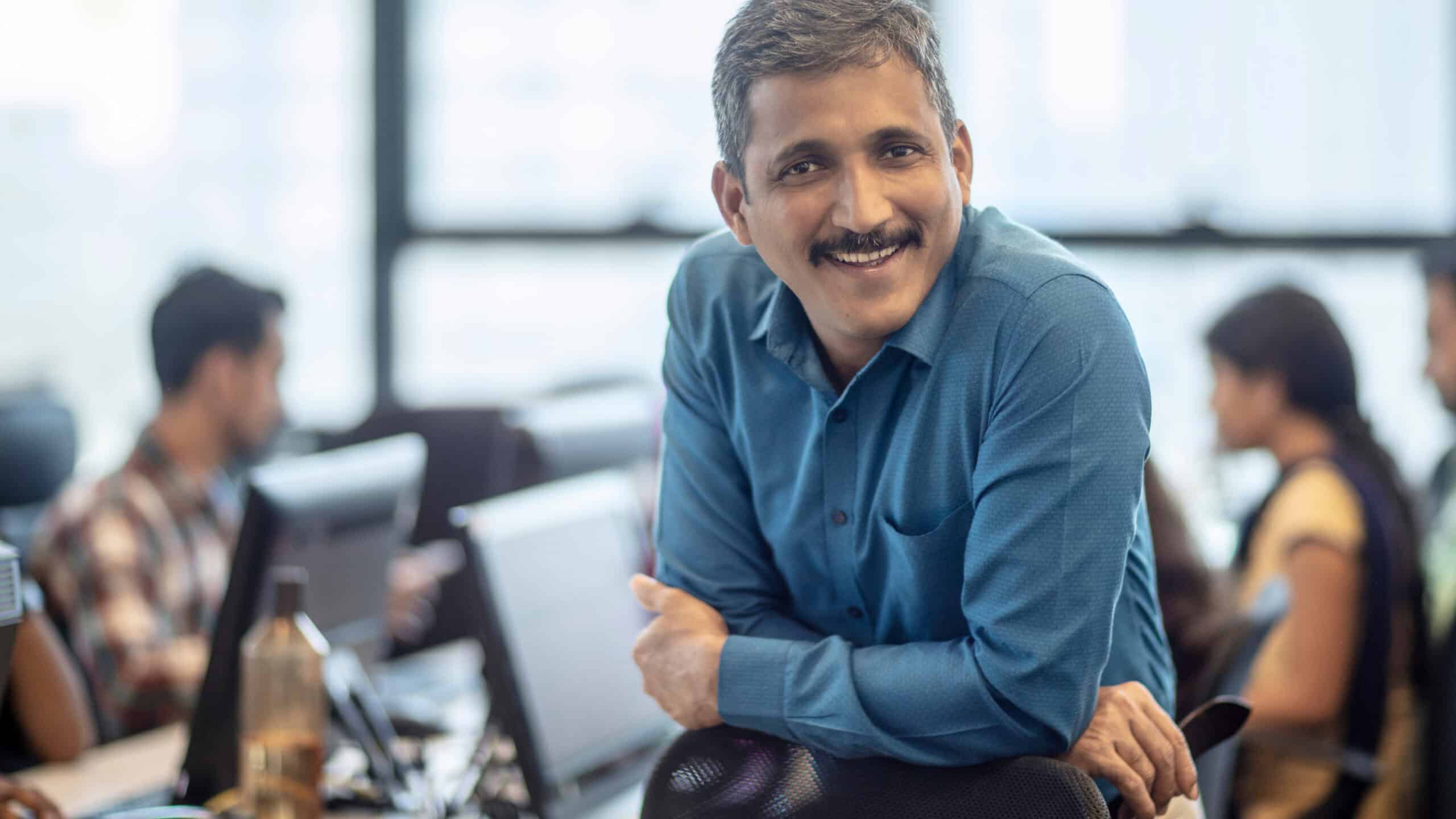 Smiling Indian manager leaning on chair.