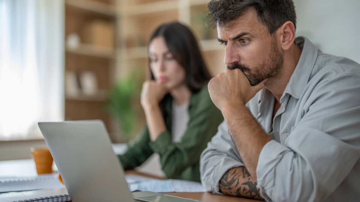 Man and woman sitting at table with the man looking a bit puzzled at his laptop.