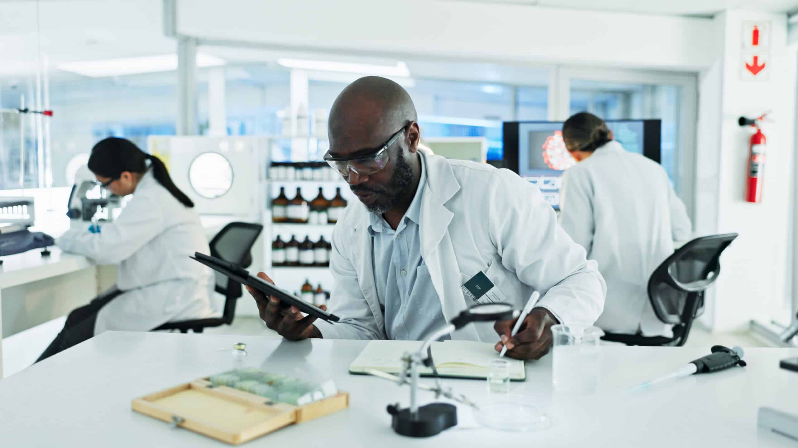 Scientist taking down notes from a tablet, with two other scientists working in the background.