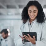 A female scientist in a laboratory setting using a tablet to review data, with a male scientist working in the background.