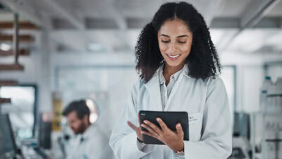 A female scientist in a laboratory setting using a tablet to review data, with a male scientist working in the background.