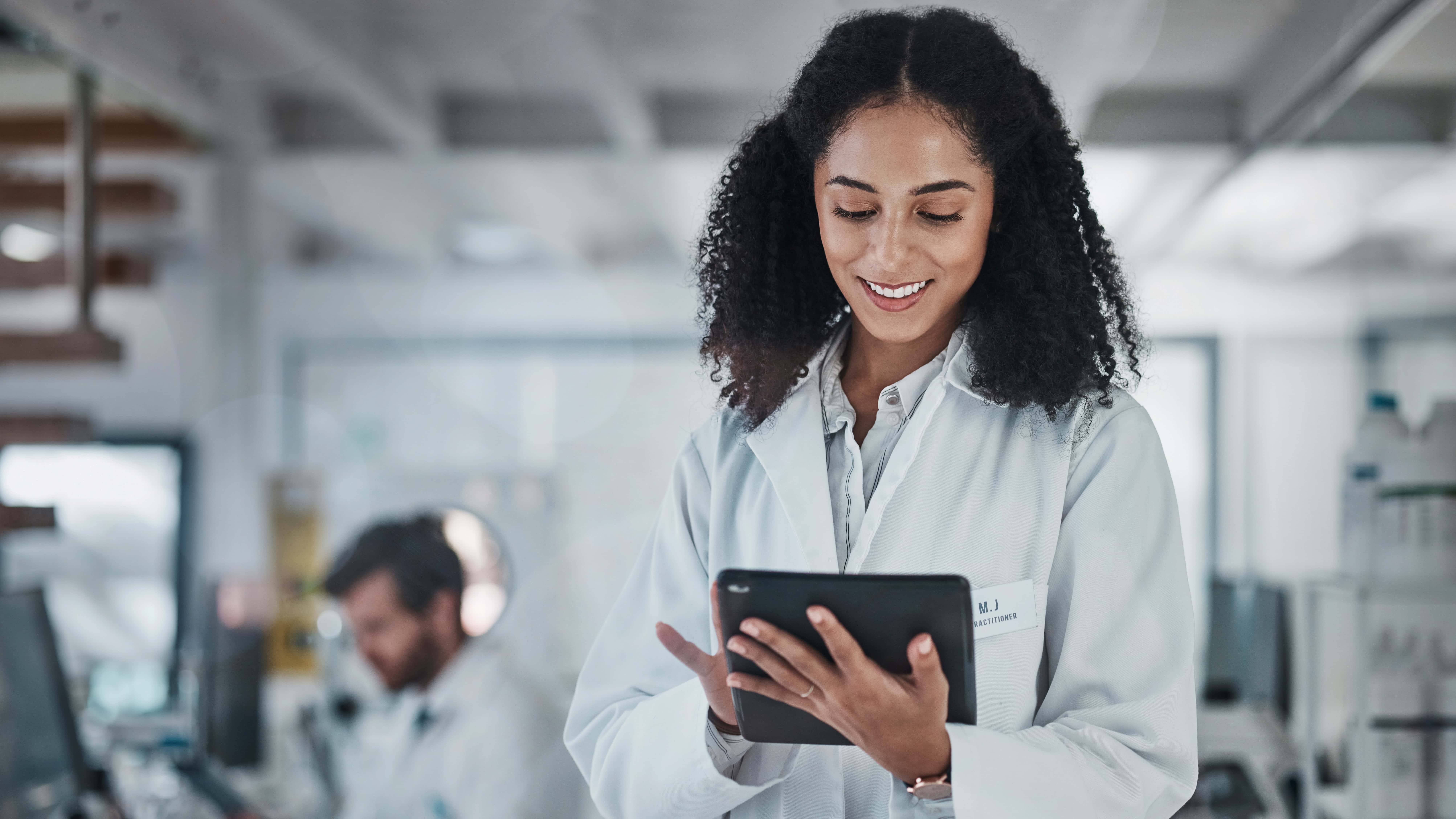 A female scientist in a laboratory setting using a tablet to review data, with a male scientist working in the background.