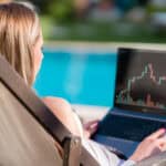 Woman sitting on a chair by the pool on her laptop, looking at a stock market chart.