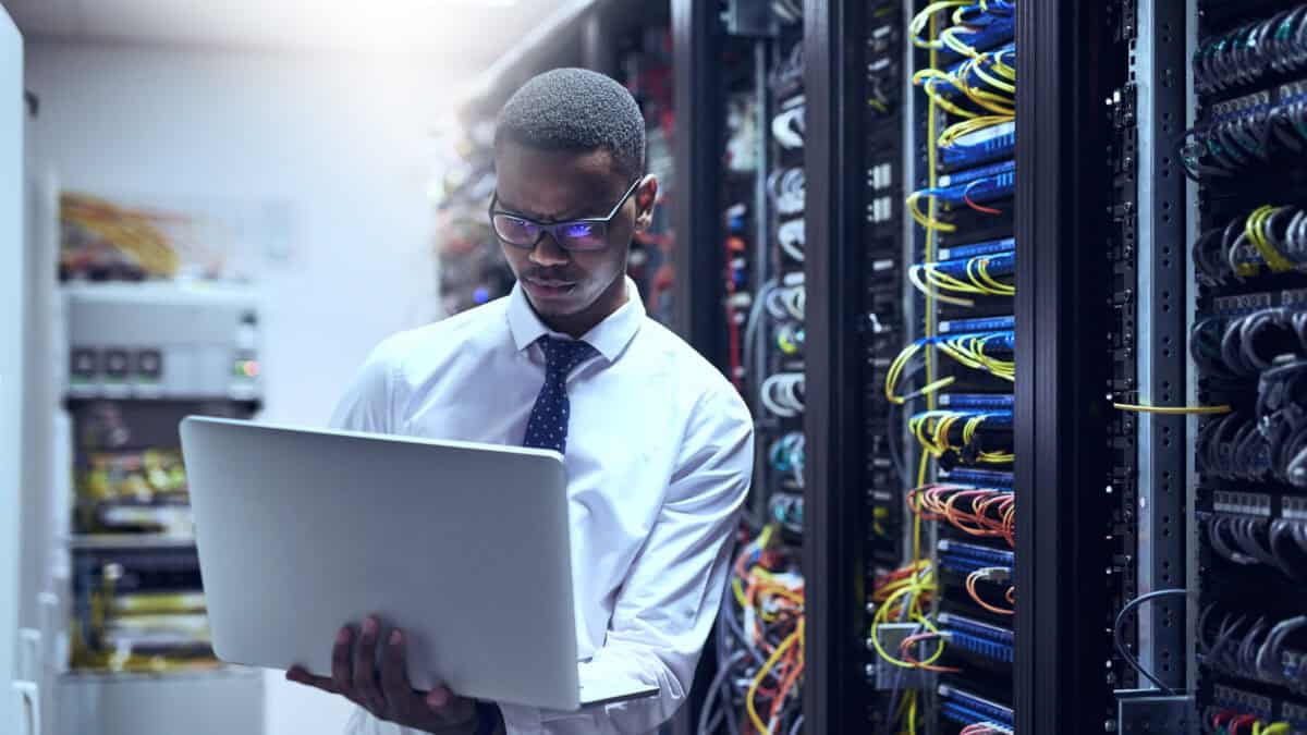 Man using a laptop next to the backside of server racks in a data centre.