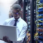 Man using a laptop next to the backside of server racks in a data centre.