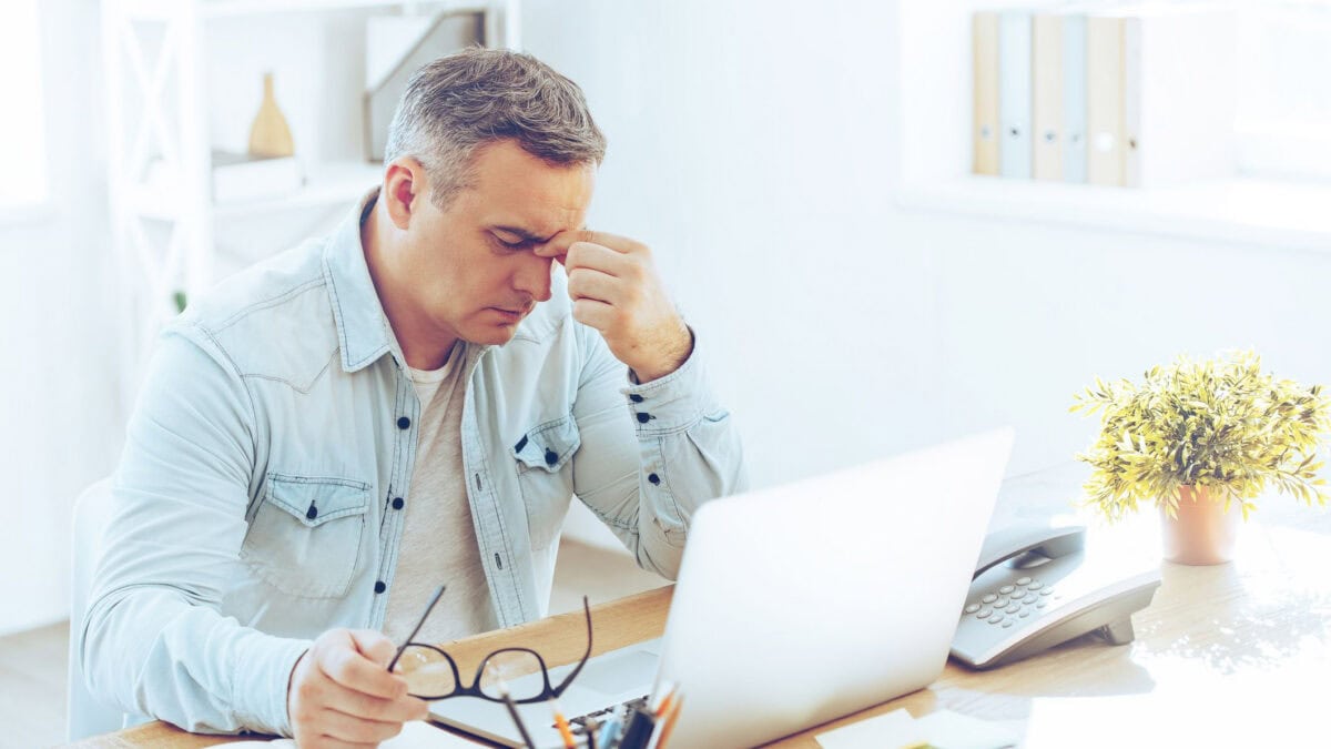 Frustrated man looking exhausted while sitting at his desk with his laptop and carrying his glasses in his hand.