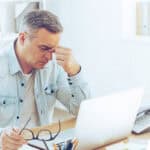 Frustrated man looking exhausted while sitting at his desk with his laptop and carrying his glasses in his hand.